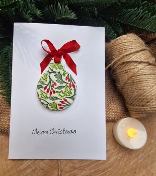 A beautiful crisp white greetings card, blank on inside. The card is adorned with a detachable clay ornament on a length of ribbon to hang on your Christmas tree. 