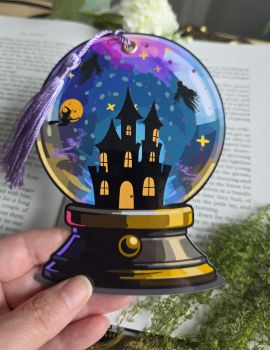 Step into a world of magic every time you open your book! Shaped like a mystical crystal ball, this handmade bookmark showcases a silhouette of a spooky haunted mansion, flying witches, and swirling wraiths set against a starry cosmic night sky. Finished with a lush silky tassel, it is the ultimate accessory for dark fantasy lovers, cosy Halloween reads, and witchy stationery collectors.