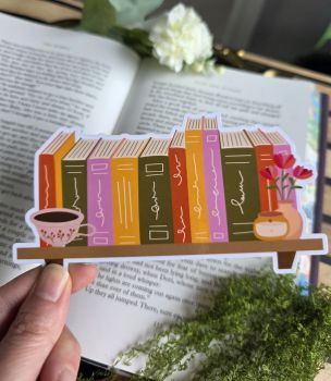 Bring the ultimate cosy reading nook straight into your favorite book! Designed in a charming bookstack shape, this die-cut bookmark showcases a wooden shelf lined with colorful spine-detailed books, a warm floral tea cup, a glowing candle, and a vase of pink blooms. It&rsquo;s the ultimate accessory for cozy fiction, contemporary reads, or anyone who dreams of having their own personal library.