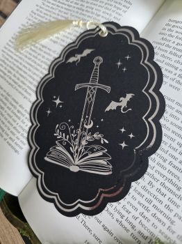 Unleash epic fantasy magic every time you open your current read! Crafted on a striking matte black card, this scalloped bookmark features a shimmering metallic silver foil design of a sword emerging from an open book, surrounded by flying dragons and celestial stars. Complete with a soft champagne-gold silky tassel, it is a luxurious, must-have keepsake for fantasy readers, dragon lore enthusiasts, and dark academia book lovers.