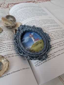 A hand-painted miniature coastal scene featuring a lighthouse, set in a handmade dark blue oval frame.