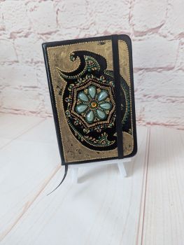 A lined, undated notebook with a hand-painted black, green and gold design, finished with decorative dot details and gemstone accents.